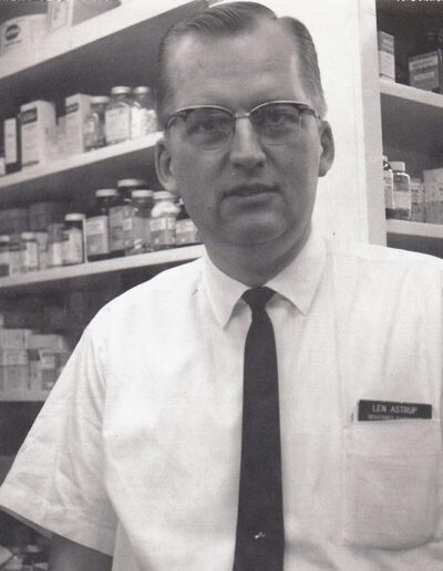 A Photo of Leonard Astrup, Wearing a Black Tie, in Front of Shelves of Medication.