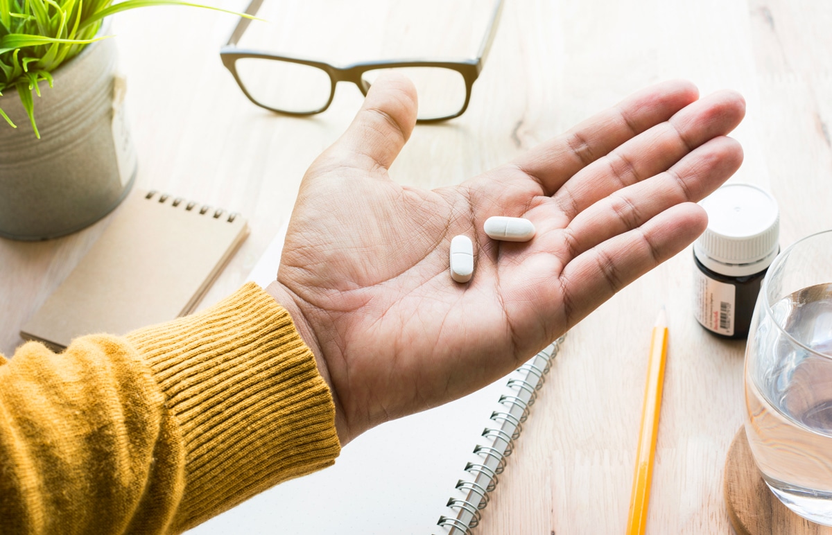 A hand with an open palm holding two pill capsules, with a desk and office materials behind the hand.